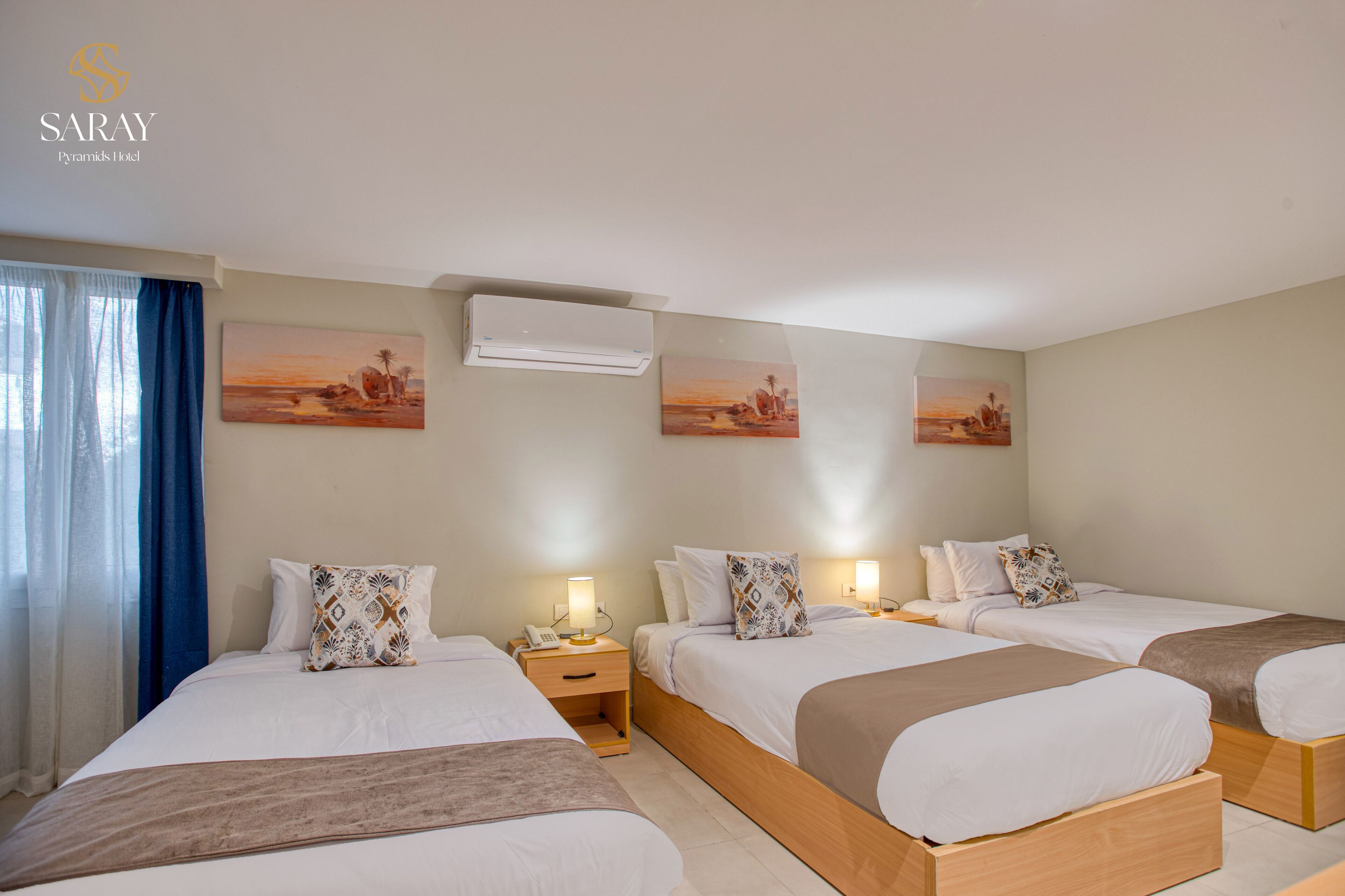 A hotel room with three single beds, a nightstand with a lamp, an air conditioner, and two wall-mounted paintings.