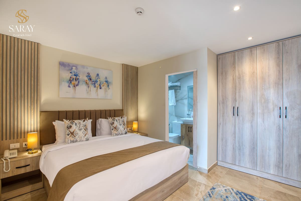 A hotel room with a large bed, a wooden headboard, a painting on the wall, a wardrobe, and a bathroom visible through an open door.
