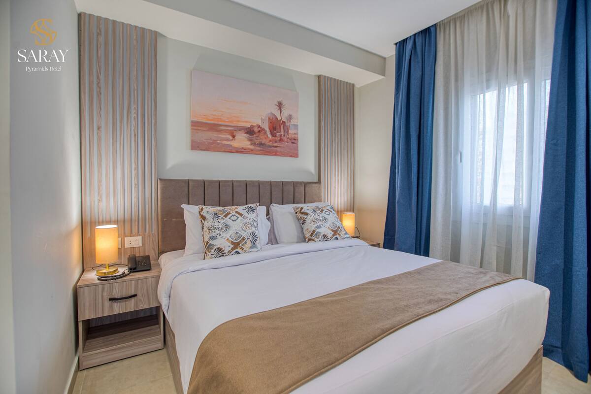 A hotel room with a large bed, two bedside tables with lamps, a painting above the bed, and a window with curtains.