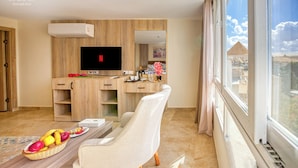 A modern hotel room with a flat-screen TV, wooden furniture, a dining area with fruits, and a view of pyramids. - Saray Pyramids & Museum View Hotel (Giza)