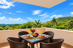 A balcony with wicker furniture, a table with breakfast items, and a scenic view of a lush landscape and ocean.