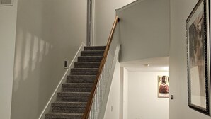 Una escalera con peldaños alfombrados y barandilla de madera, que conduce a un rellano con una puerta y un cuadro en la pared.
