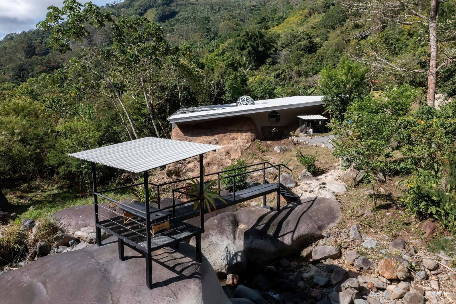 Un espacio de estar al aire libre rústico con techo de metal y bancos, rodeado de formaciones rocosas y vegetación.