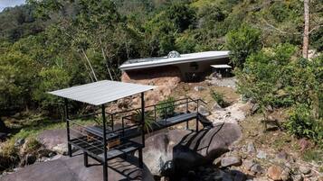 Un espacio de estar al aire libre rústico con techo de metal y bancos, rodeado de formaciones rocosas y vegetación.