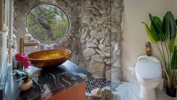 A bathroom with a stone wall, a round window, a large bowl sink, a toilet, and a potted plant.