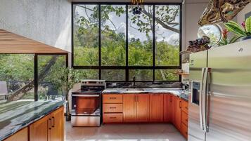 A modern kitchen with wooden cabinets, a stainless steel refrigerator, and a large window offering a view of greenery.