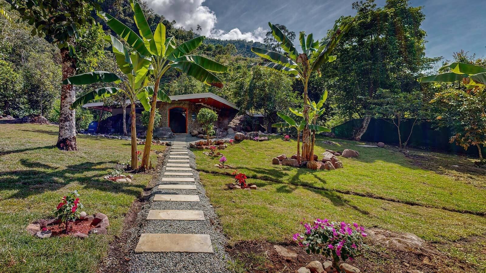 Un sendero conduce a una construcción rústica rodeada de abundante vegetación y palmeras de plátano.
