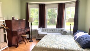 Ein Schlafzimmer mit einem Bett, einem Klavier, einer Couch und zwei mit Vorhängen versehenen Fenstern.
