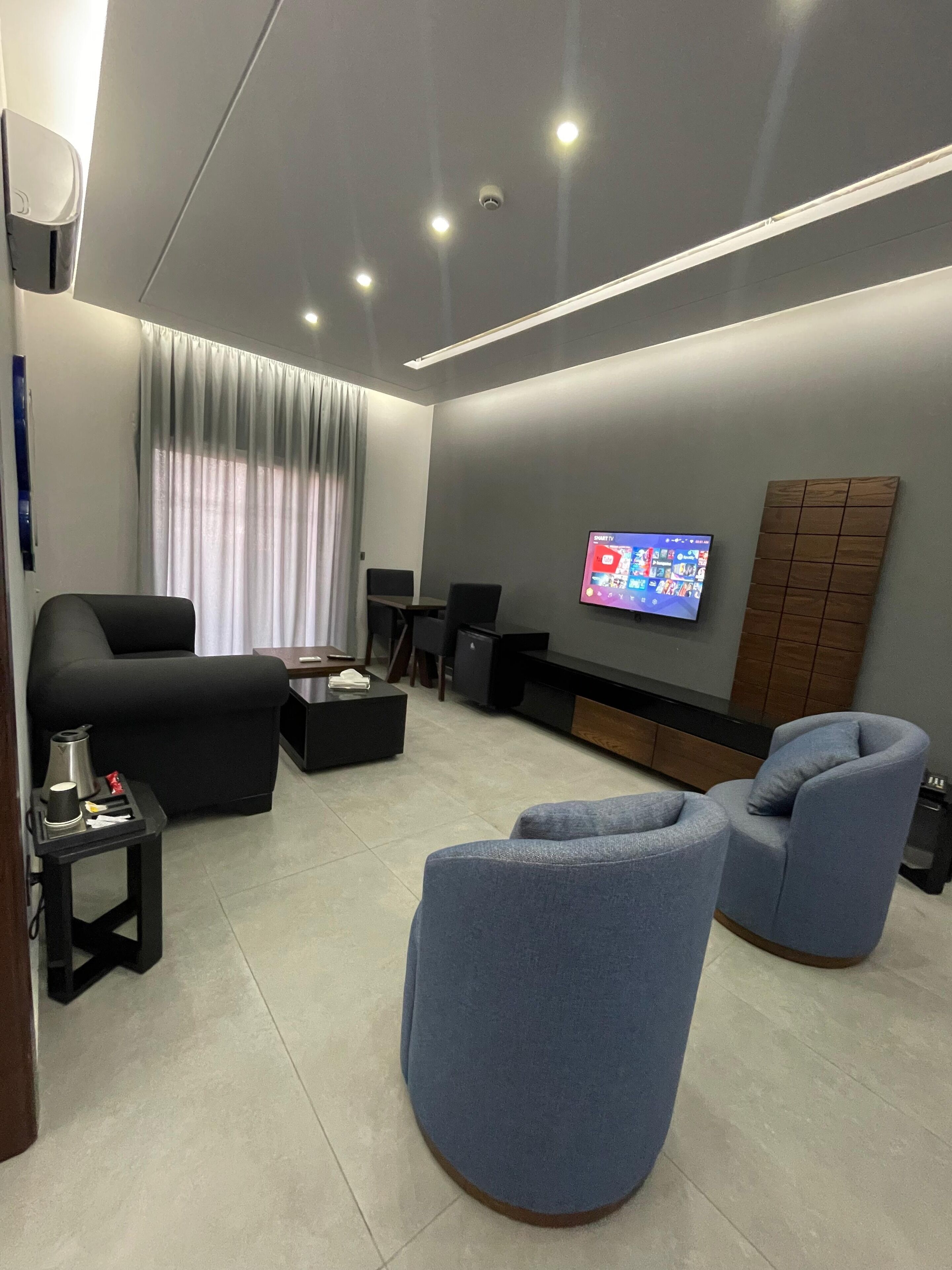 Una sala de estar moderna con un televisor de pantalla plana montado en la pared, un sofá, dos sillones, una mesa de centro y una mesita auxiliar con una tetera y una taza.