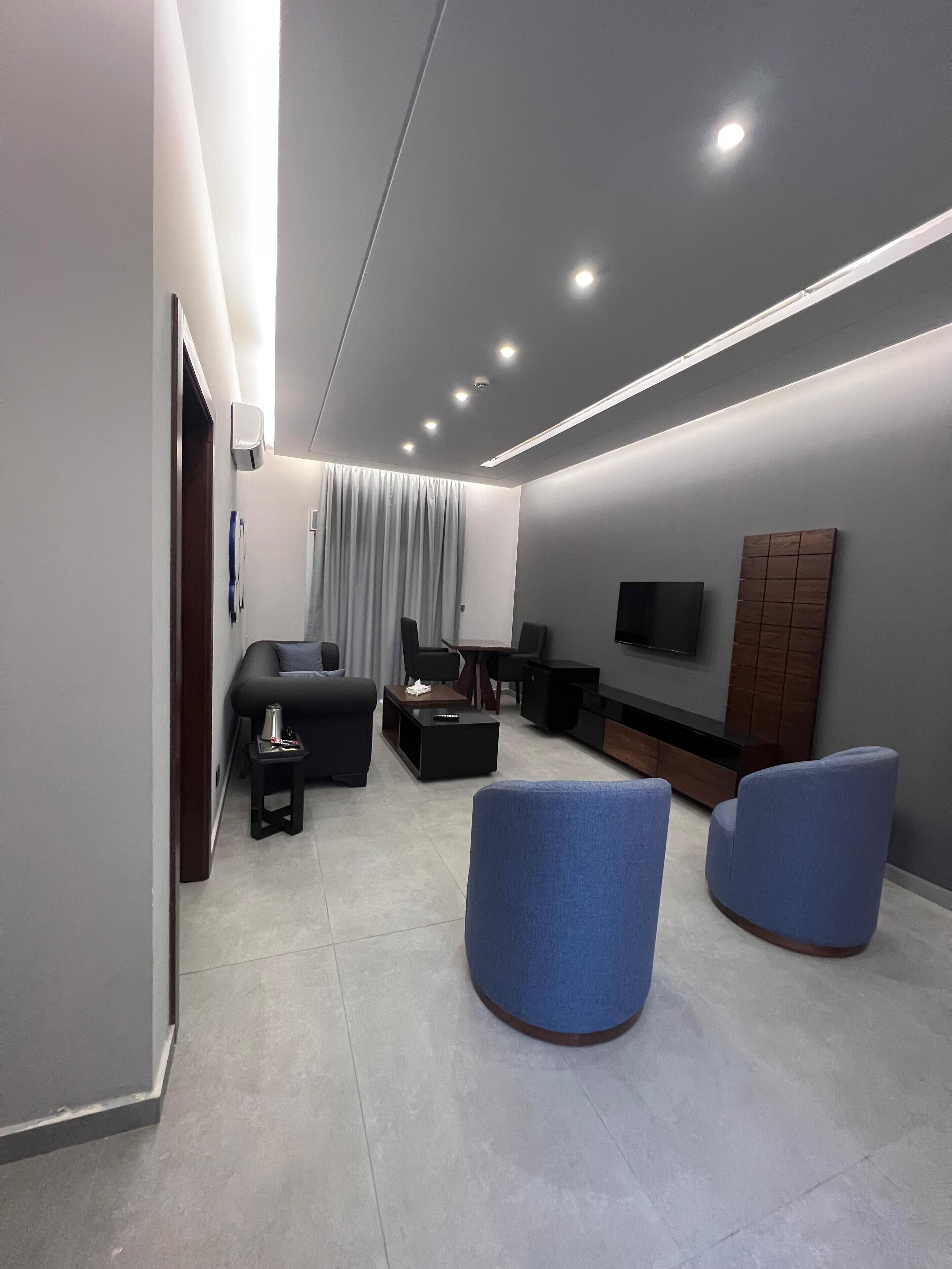 A modern living room with a flat-screen TV, dark sofas, a coffee table, and a blue accent chair.