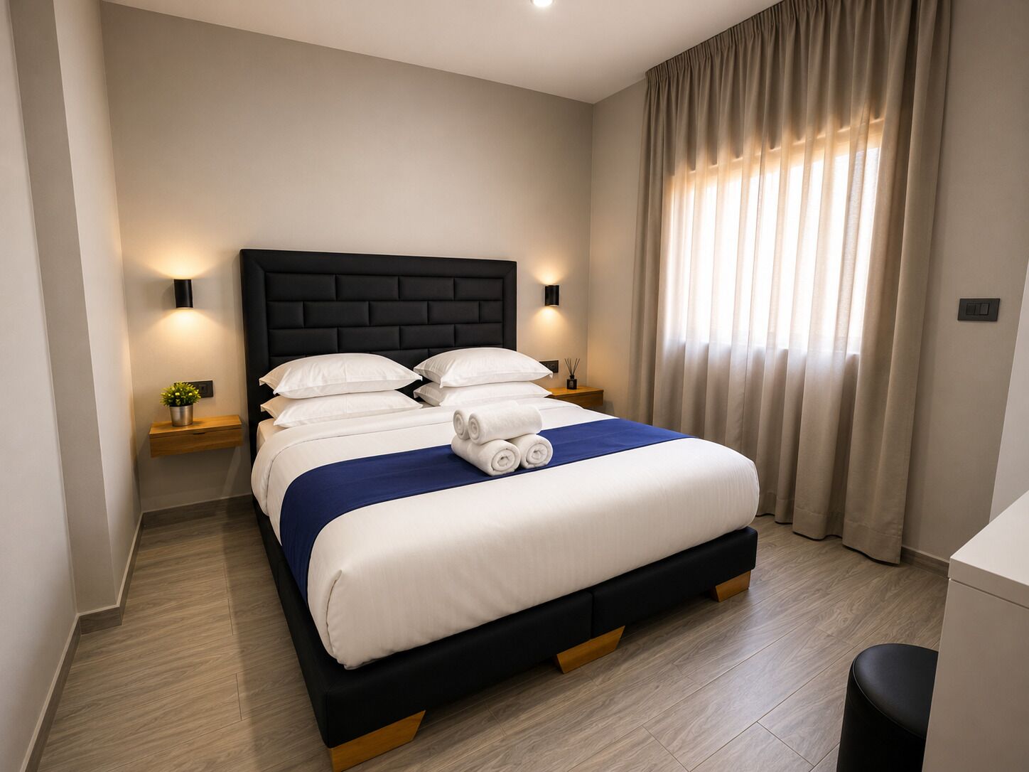 Et moderne hotelværelse med en stor seng, sengeborde med lamper, et lille bord med en plante og et vindue med gardiner.
