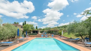 Una piscina rectangular con sillas reclinables y sombrillas azules, rodeada de vegetación y un cielo despejado.