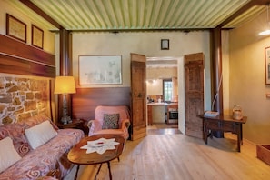 Una stanza accogliente con una parete in pietra, un divano decorato, un tavolo in legno e una zona cucina visibile attraverso una porta aperta.