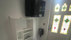 A room with a white desk, a laptop, a bottle, and a cup. There is a TV mounted on the wall and a door with stained glass windows.