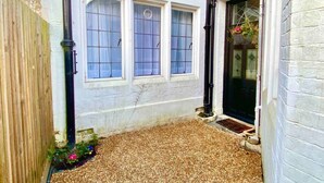 A small, enclosed garden area with a gravel path, a wooden fence, and a white building with three windows and a black door.