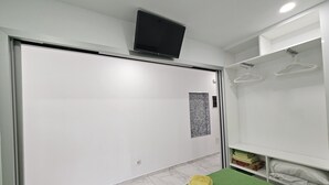 Una camera d'albergo con un letto grande, un tavolino con un asciugamano verde, un armadio con abiti appesi e un televisore a parete.