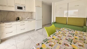 Cucina compatta con mobili bianchi, un forno a microonde e un frigorifero. C'è un divano verde e un tavolo con tovaglia floreale.