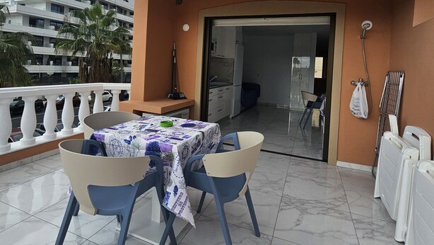 En balkon med et bord og stole, udsigt til en bygning, og en dør der fører ind.