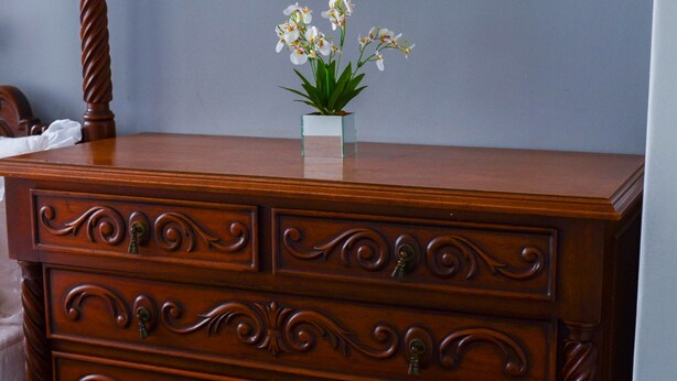 A wooden dresser with ornate carvings, a framed picture on the wall, and a small vase with flowers on top. - Refuge for your family (Centro Guarapari)