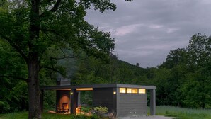 Un chalet moderne doté d’une cheminée, entouré d’une végétation luxuriante et situé au pied d’une colline boisée.