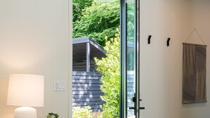 A modern entryway with a glass door leading to a garden, a round table with a lamp and a plant, and a wooden stool.
