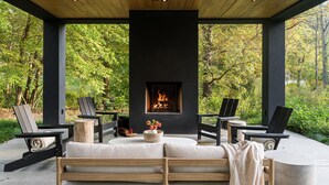 An outdoor seating area with a fireplace, surrounded by chairs and a bench, set against a backdrop of trees.