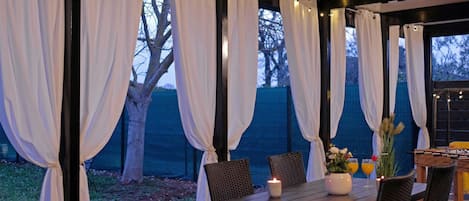 An outdoor dining area with a table and chairs, draped white curtains, and string lights.