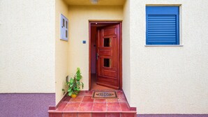 Una porta de fusta amb un felpú de benvinguda, una planta en test i una finestra amb persianes blaves a la façana d'un edifici.