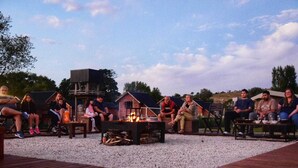 A group of people seated around a fire pit on a gravel area with wooden decking, with buildings in the background.