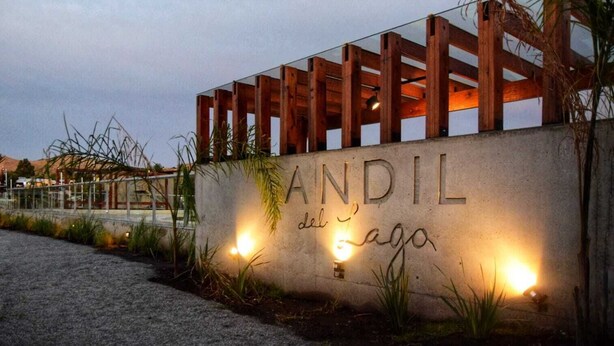 A concrete wall with the name 'ANDIL del lago' illuminated by lights, surrounded by wooden structures and plants.