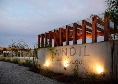A concrete wall with the name 'ANDIL del lago' illuminated by lights, surrounded by wooden structures and plants.. Tandil del Lago