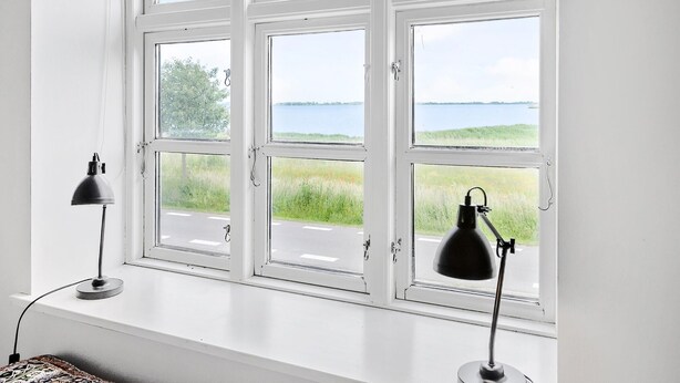 Ein Zimmer mit großem Fenster mit Blick auf eine Wiese und ein Gewässer. Zwei Schreibtischlampen stehen auf der Fensterbank.