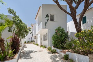 A narrow pathway flanked by white buildings with shutters, a street lamp, and various plants.