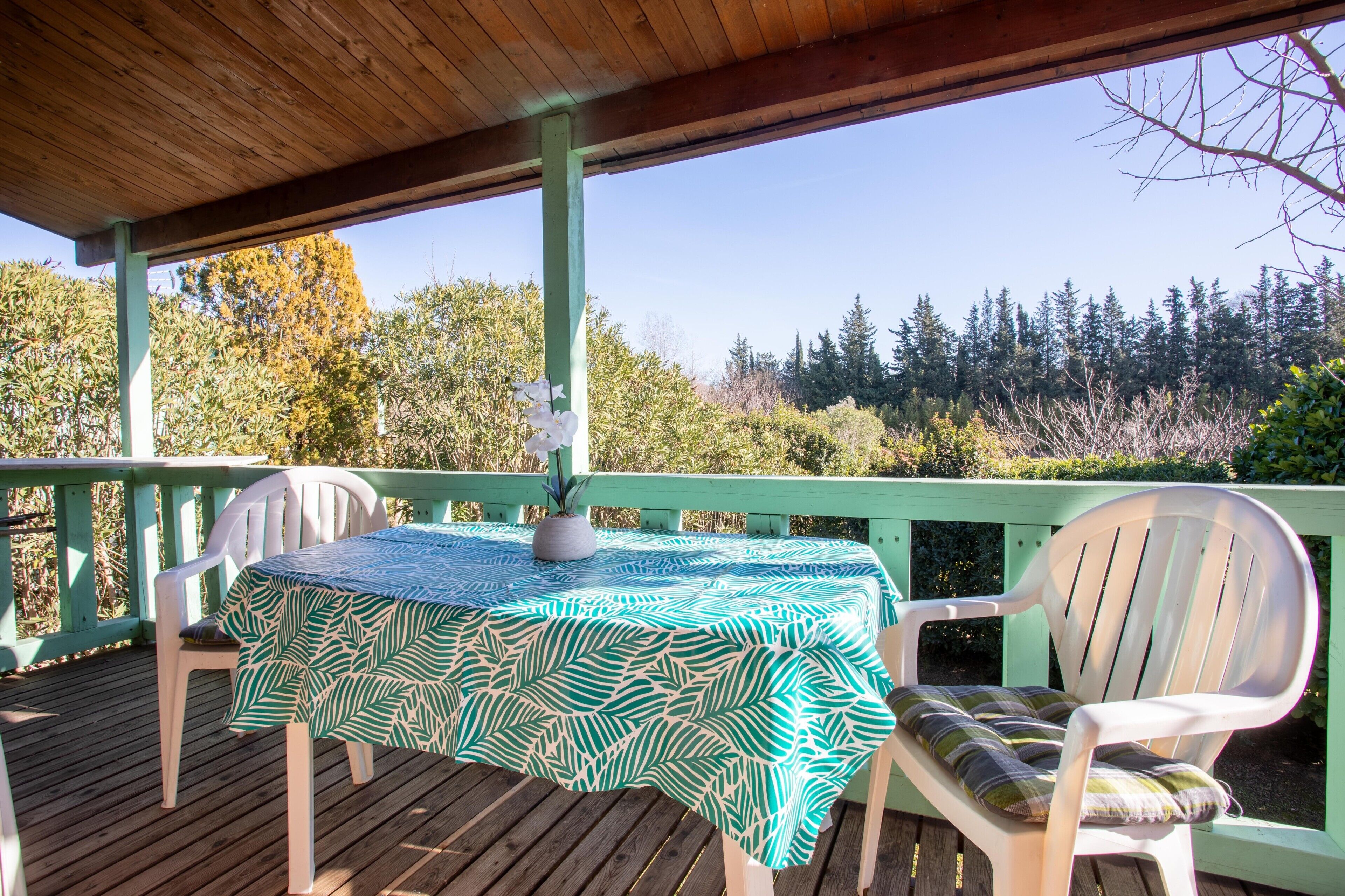 A covered outdoor deck with a table and chairs, surrounded by greenery.