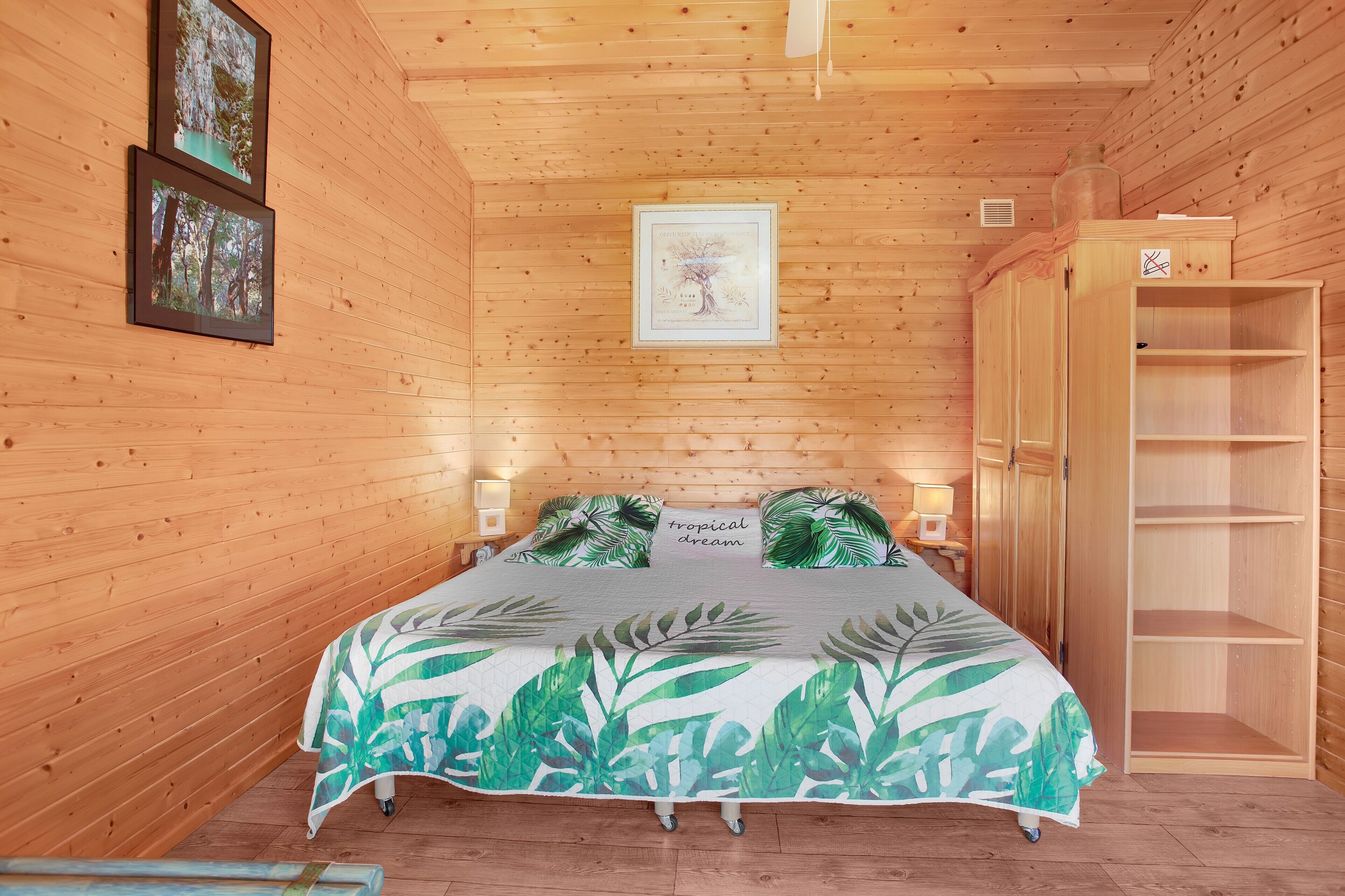 A wooden cabin bedroom with a bed, framed pictures, a nightstand, and a wardrobe.