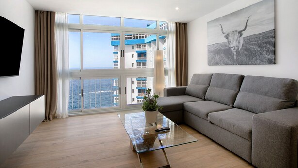 Un salon moderne avec un canapé, une table basse en verre, un téléviseur fixé au mur et une grande fenêtre avec des rideaux.