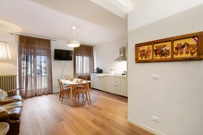 A modern living room with a sofa, dining table, and chairs, a kitchen area with cabinets and a stove, and a wall-mounted TV.