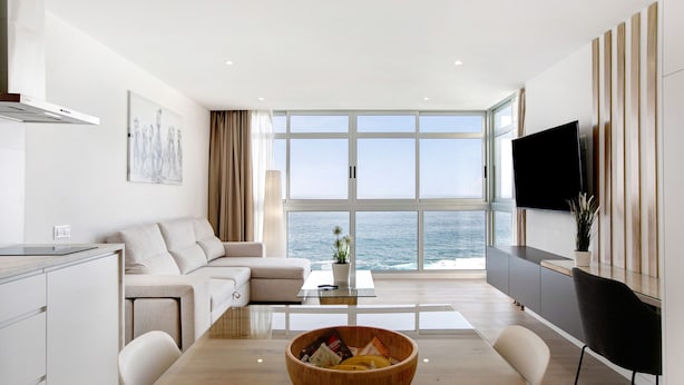 A modern living room with a large window, a wooden dining table, a sofa, and a TV mounted on the wall. - Holiday Apartment 'Luxury Loft El Roque 1' with Sea View and Wi-Fi (Tacoronte)