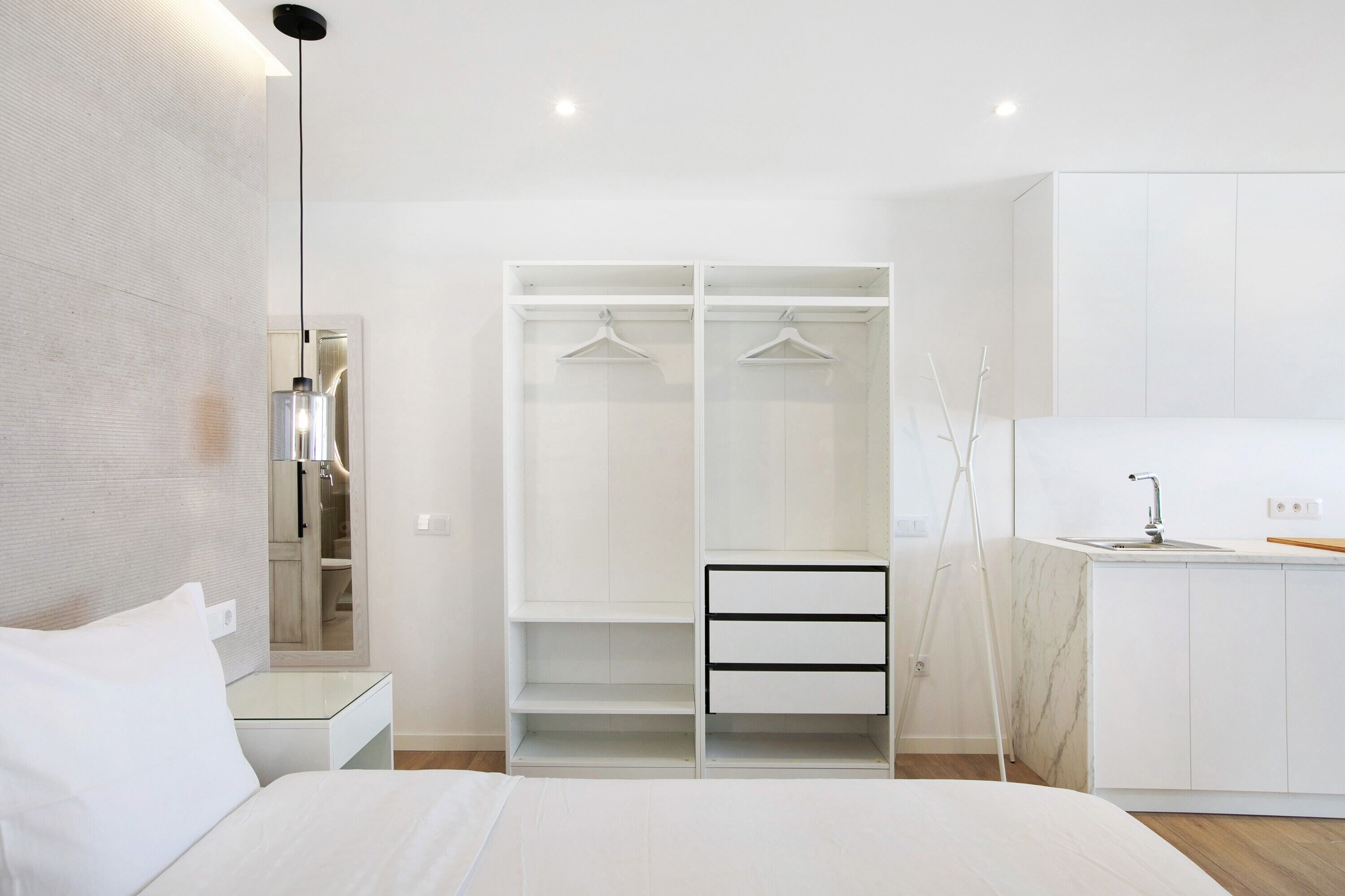 A modern bedroom with a bed, a white wardrobe with hanging space, a small bedside table, and a mirror.