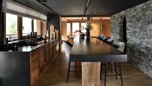 A modern kitchen with a large island, wooden cabinets, and a stone accent wall. - Chalet Enigma – Luxury at the Foot of the Slopes for 17 Guests with Spa (LA CLUSAZ)