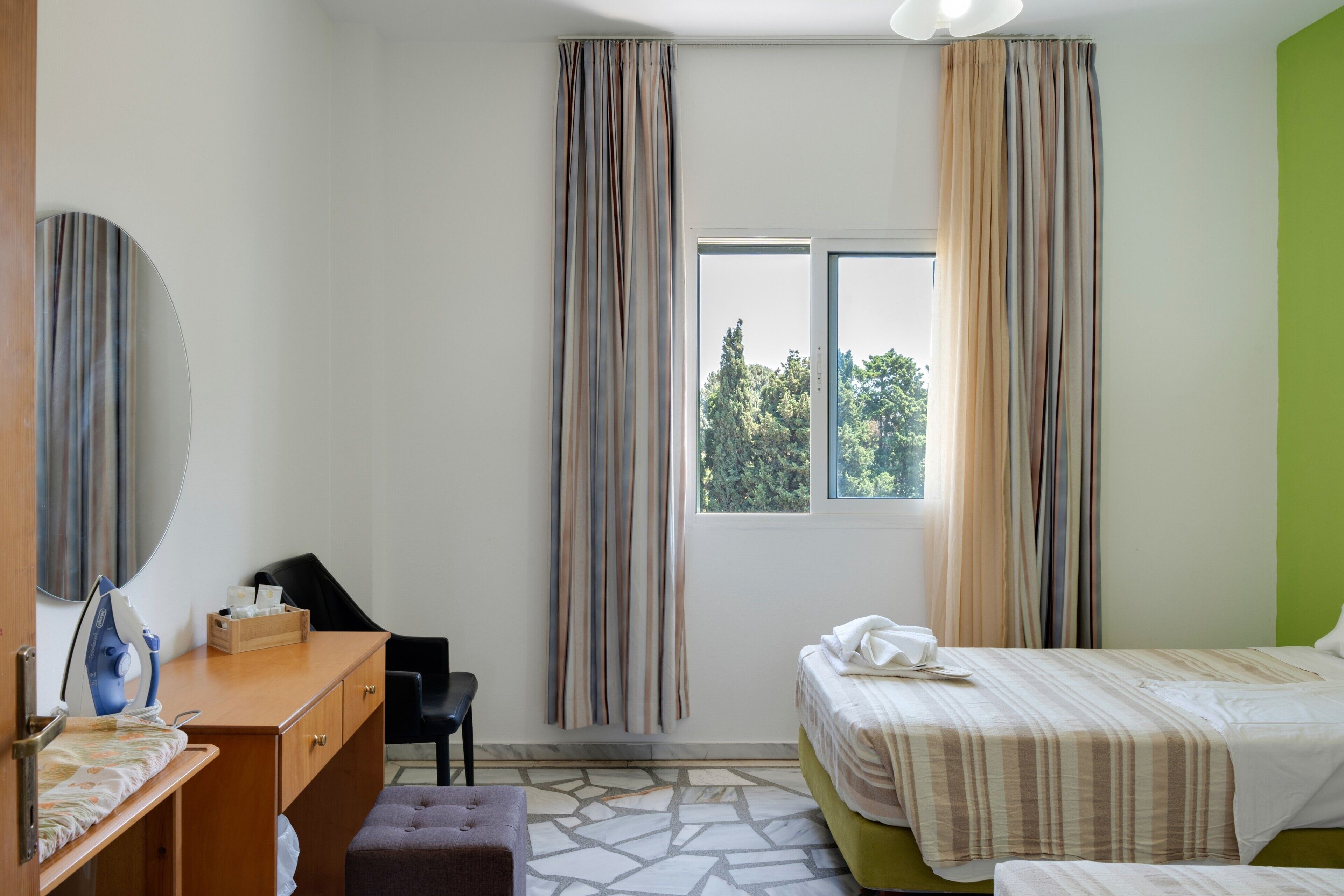 Ein Hotelzimmer mit Bett, Schreibtisch inklusive Bügeleisen, Stuhl, Fenster mit Vorhängen und Blick auf Bäume.
