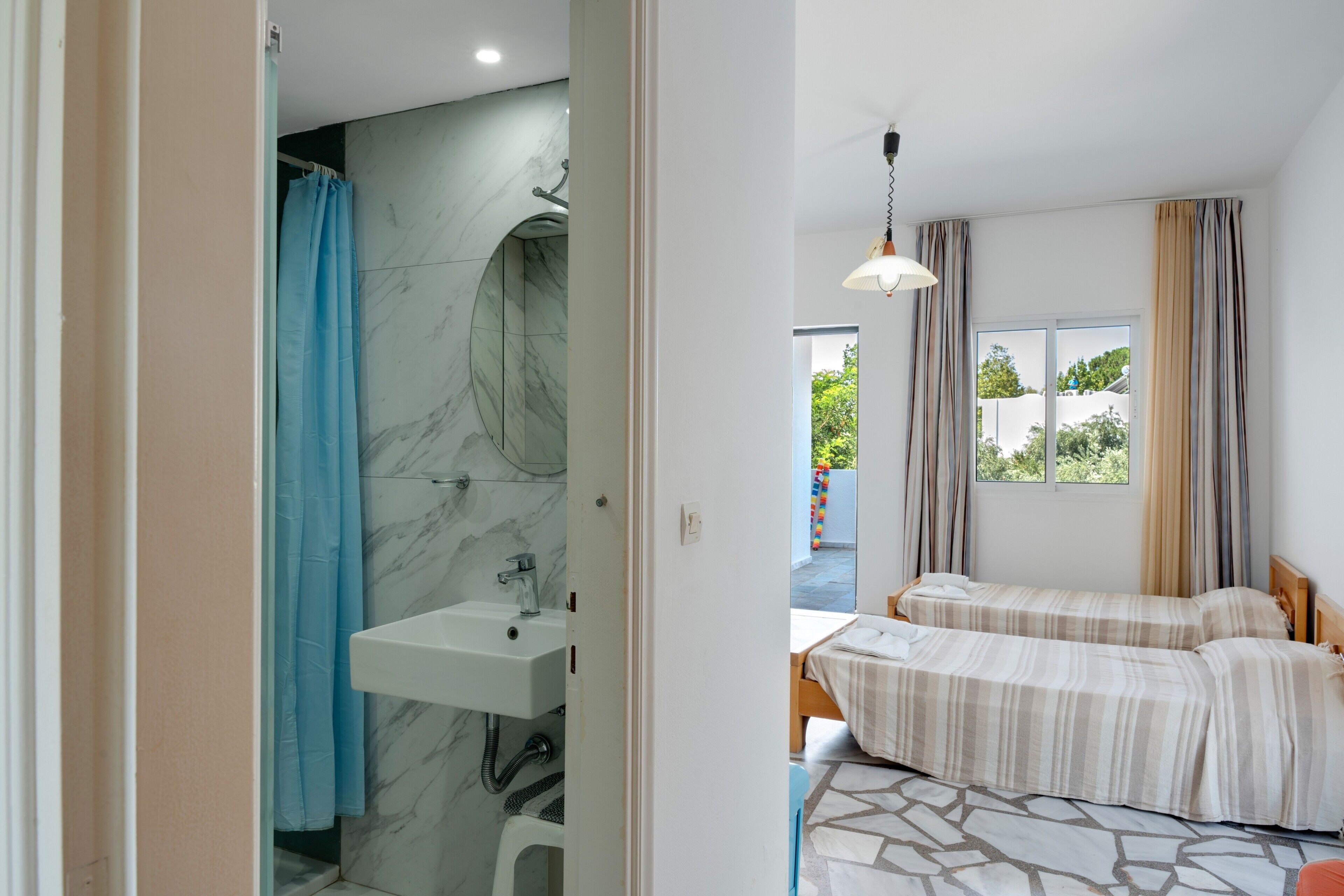 A room with a bathroom, a sink, a shower, two beds, a window, and a hanging light fixture.