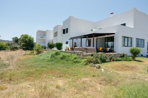 Un edificio moderno bianco con una zona salotto esterna coperta, pareti in pietra e un paesaggio verdeggiante.