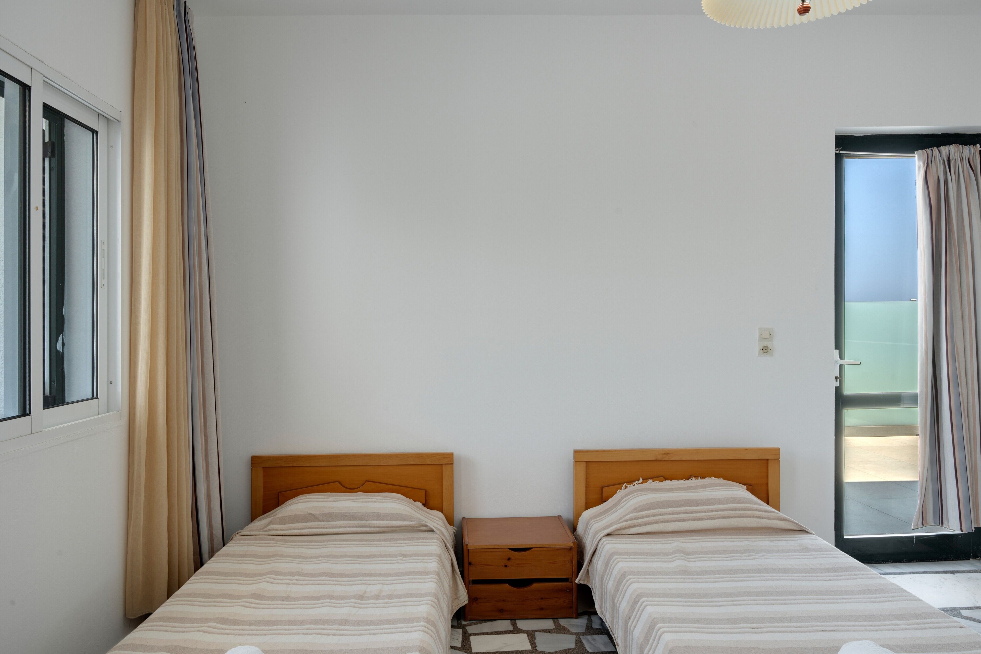 A room with two beds, a wooden headboard, a small wooden nightstand, and a sliding glass door leading to an outdoor area.