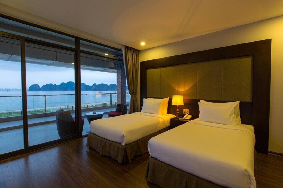 A hotel room with two beds, a large window with a view of the sea, a small table, and a chair.