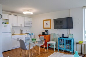 A compact kitchen with white cabinets, a microwave, and a coffee maker. There is a dining area with a glass table and chairs. A television is mounted on the wall, and a bookshelf is visible. - Fort Myers Beach Studio w/ Balcony & Views! (Fort Myers Beach)