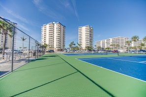 A tennis court with a chain-link fence, surrounded by tall buildings and palm trees.