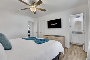 A bedroom with a bed, a ceiling fan, a TV mounted on the wall, a dresser, and a bathroom visible through an open door. - New W/ Heated SaltWater Pool! Ideal for 2 Families (Cape Coral)