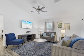 A modern living room with a flat-screen TV, a ceiling fan, and a large window with curtains. - New W/ Heated SaltWater Pool! Ideal for 2 Families (Cape Coral)