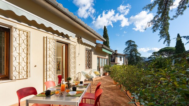 A patio with a table set for dining, surrounded by red chairs, and a view of a building with decorative windows and a tiled roof.